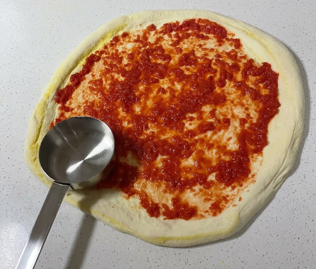 A metal ladle spreading vibrant red tomato sauce in a circular pattern over the surface of raw pizza dough.