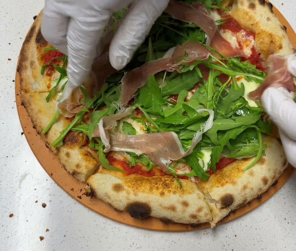 Gloved hands layering translucent slices of pink Parma ham over a bed of green arugula on a baked pizza crust.