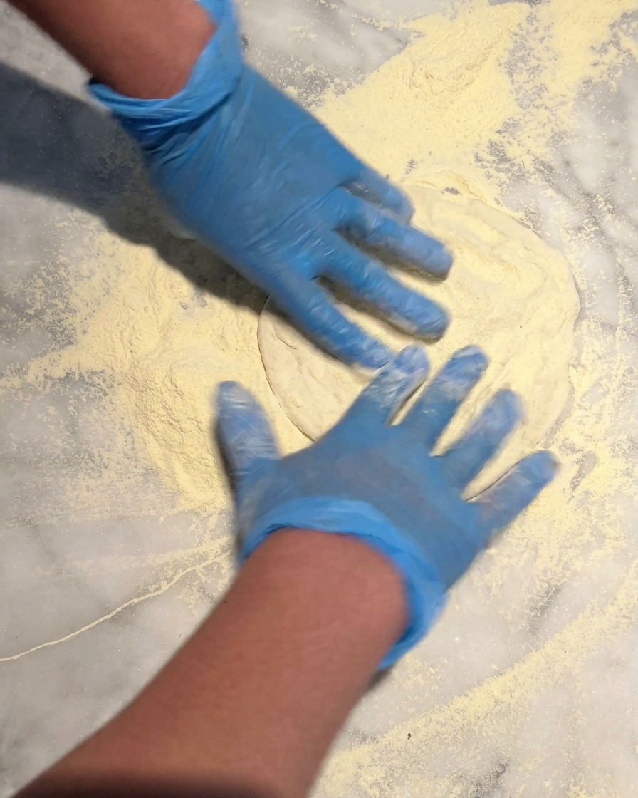 Hands in blue gloves pressing a round pizza dough ball into a thick bed of semolina flour on a marble countertop.