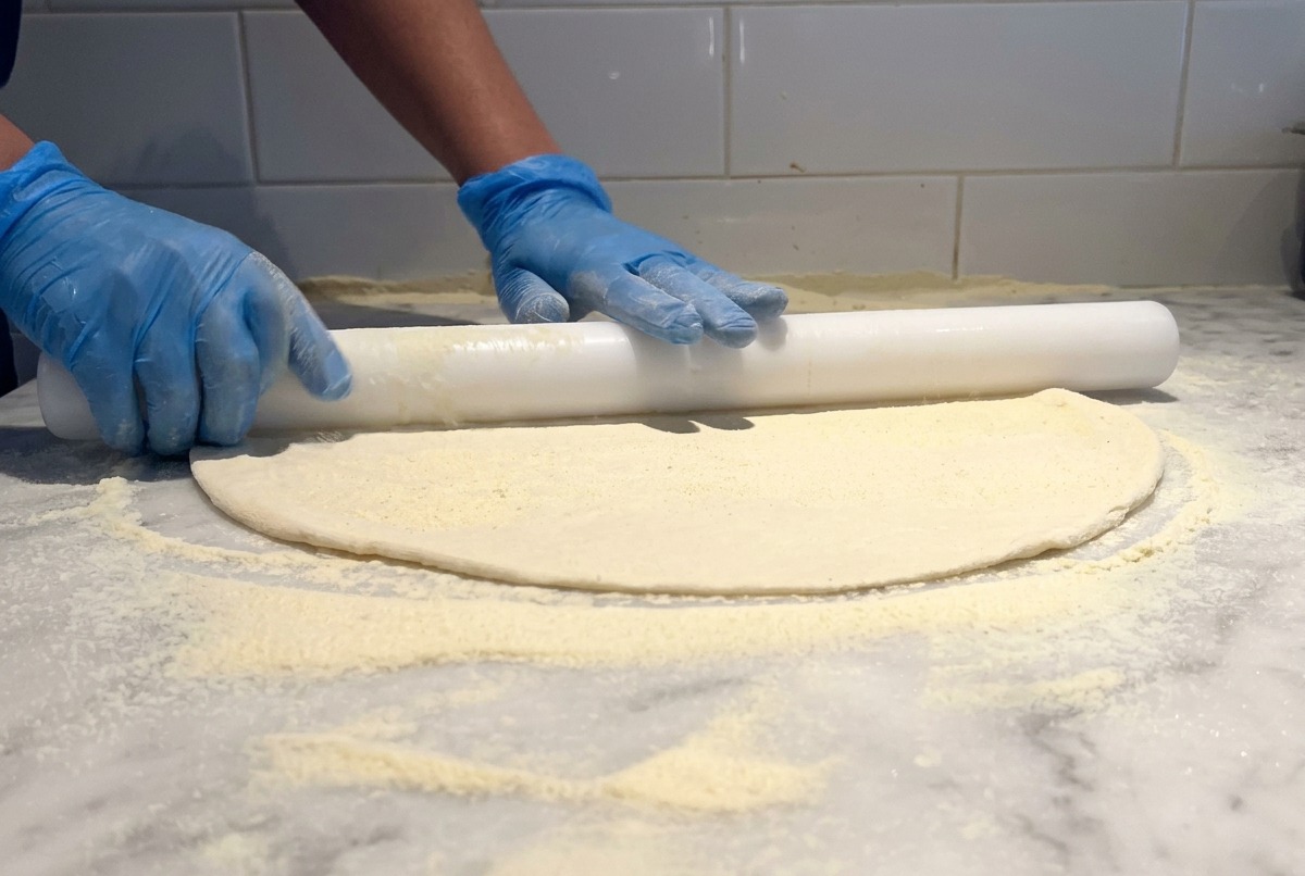 A white rolling pin being used to stretch out a thin pizza dough base on a marble table dusted with flour.