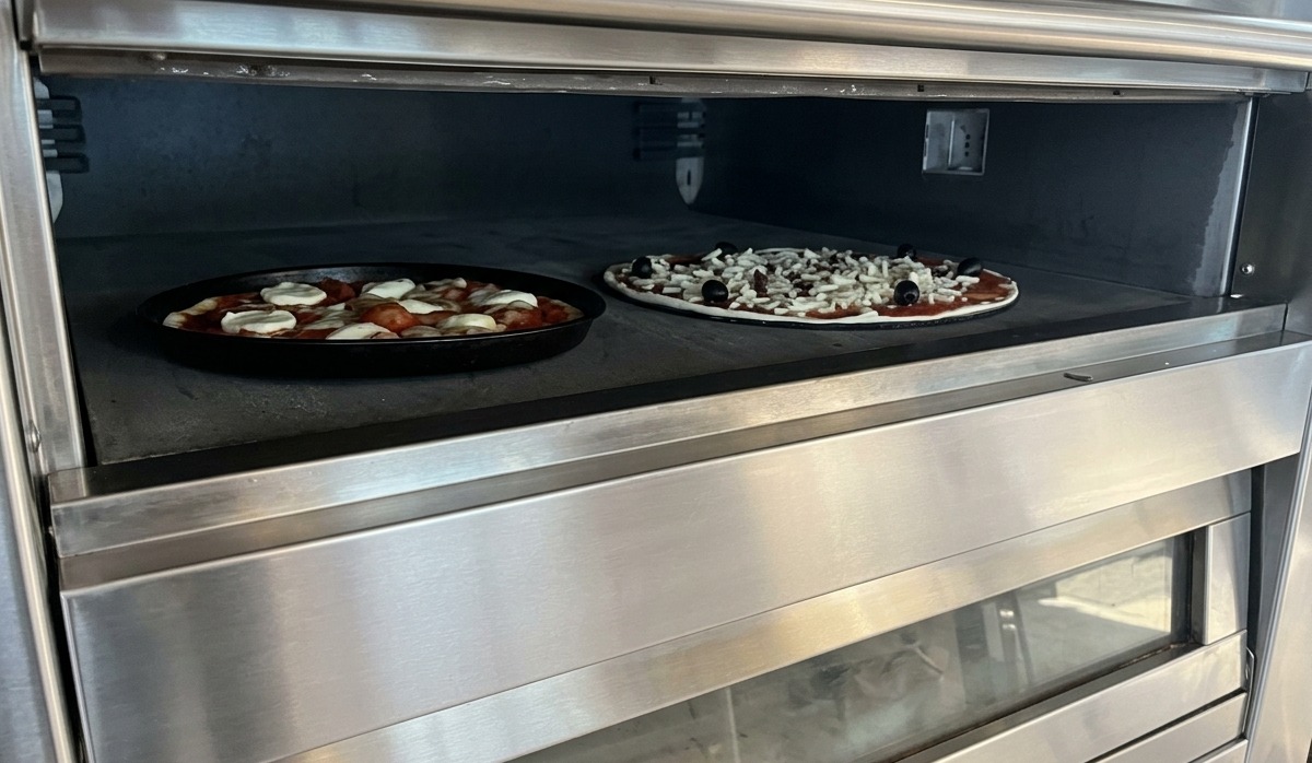 Two pizzas baking on a stone surface inside a professional stainless steel deck oven.