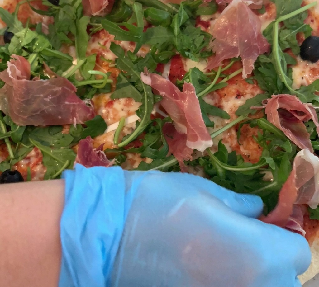 A gloved hand carefully placing thin slices of prosciutto over a pizza topped with fresh arugula.