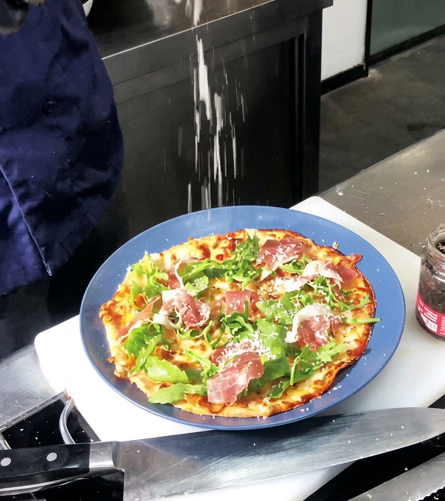 Fine white Parmesan powder being sprinkled over a baked pizza topped with green arugula and red ham slices.