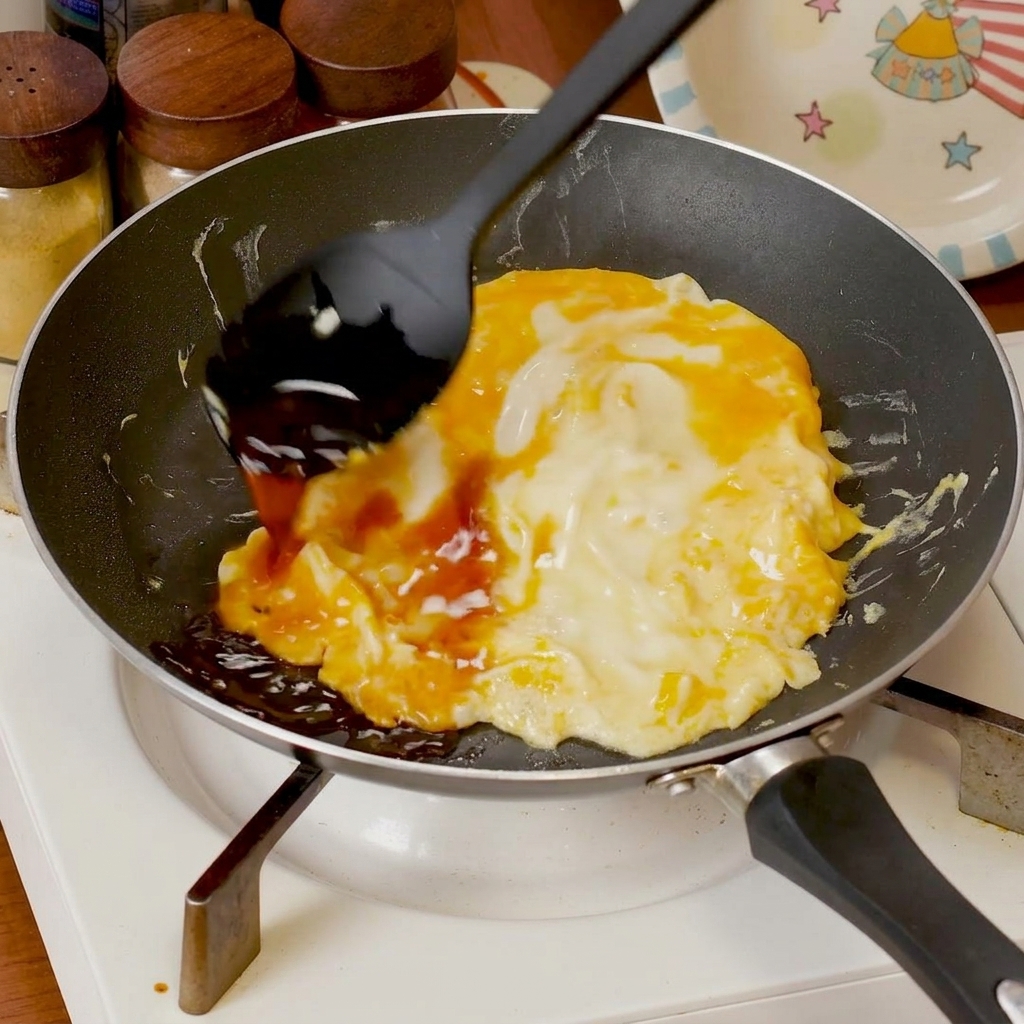 Savory sauce being poured over soft-scrambled eggs cooking in a pan.