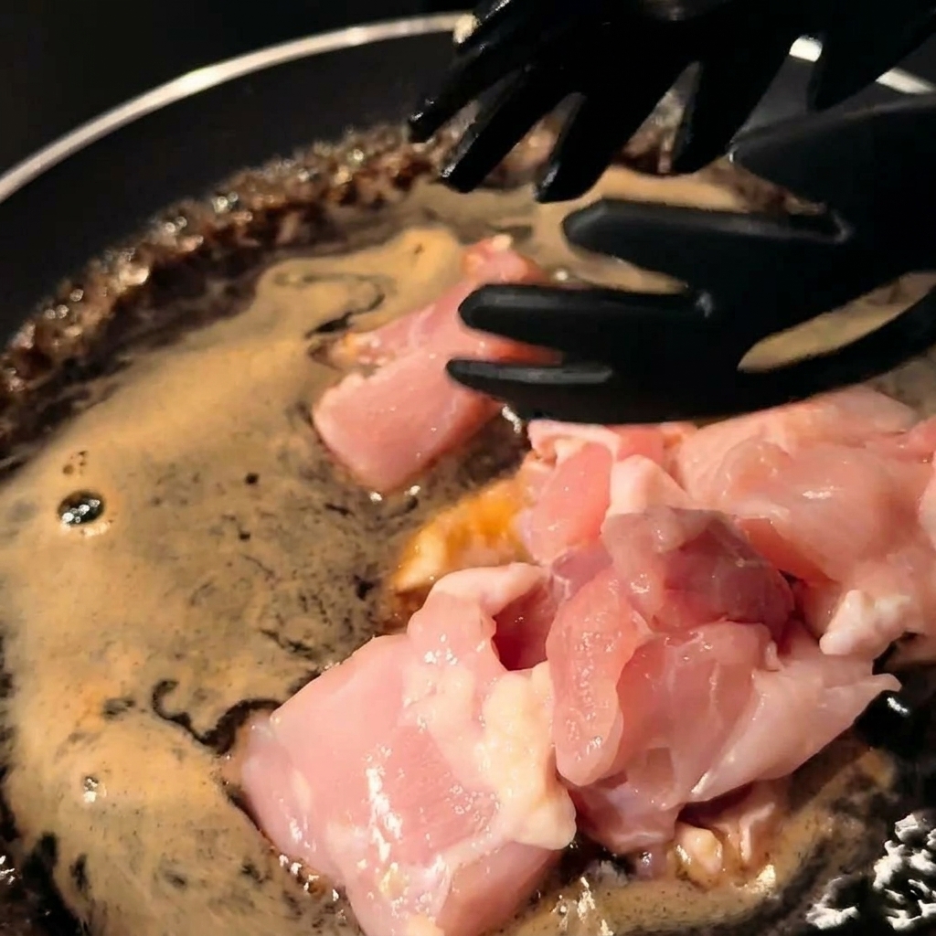 Chicken pieces and sliced onions being stirred into the sauce in a pan.