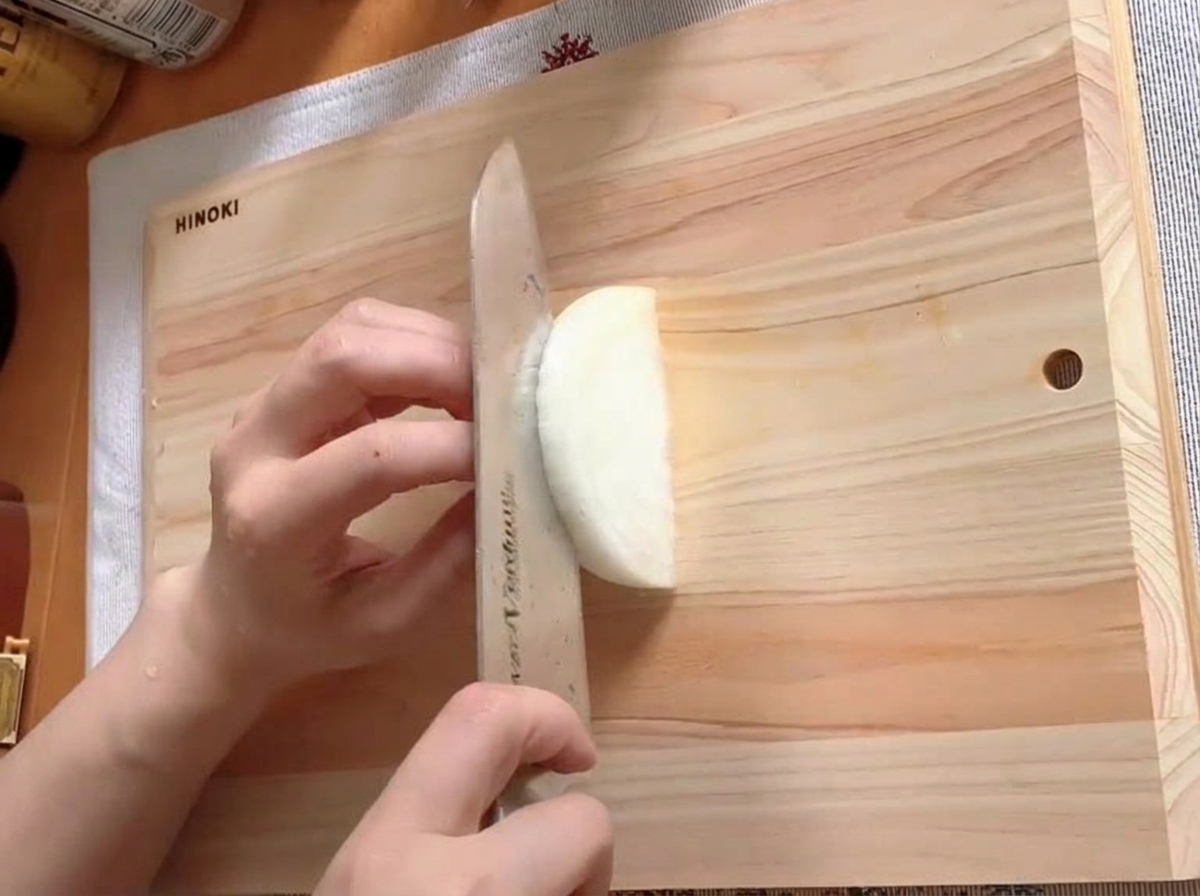 Half an onion being thinly sliced on a wooden cutting board.