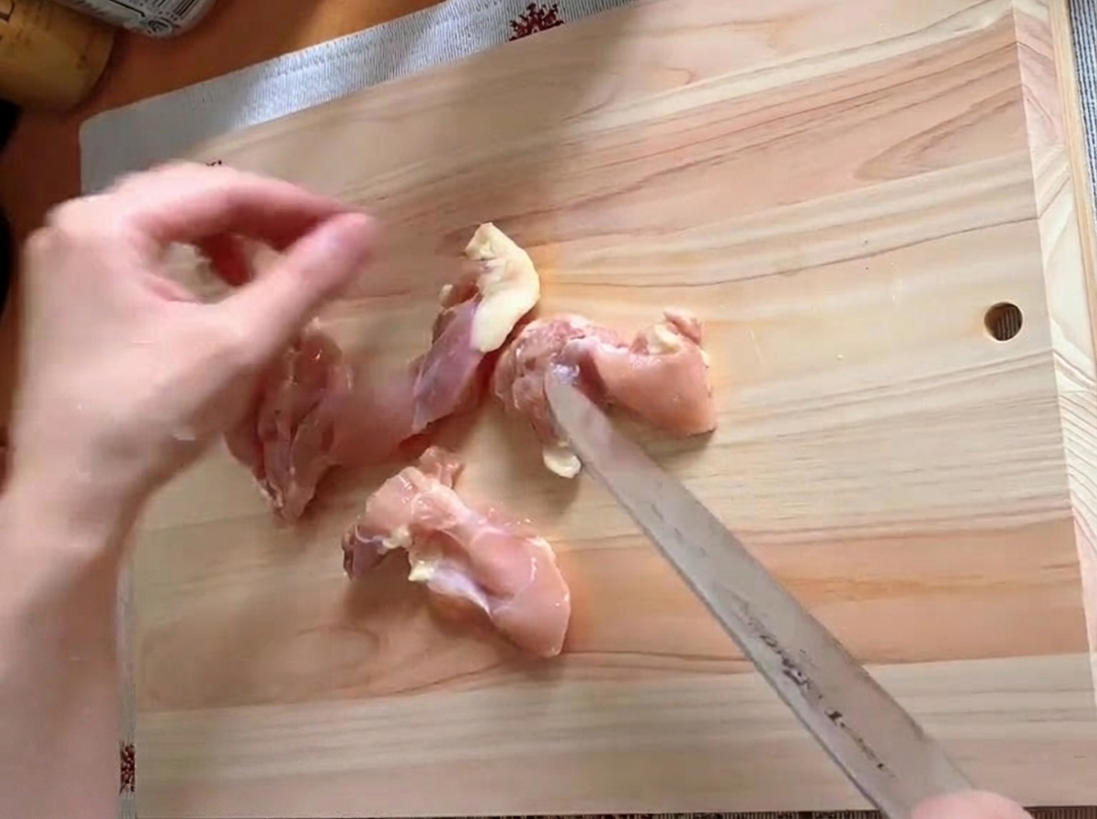 Chicken thigh being trimmed and cut into pieces on a cutting board.