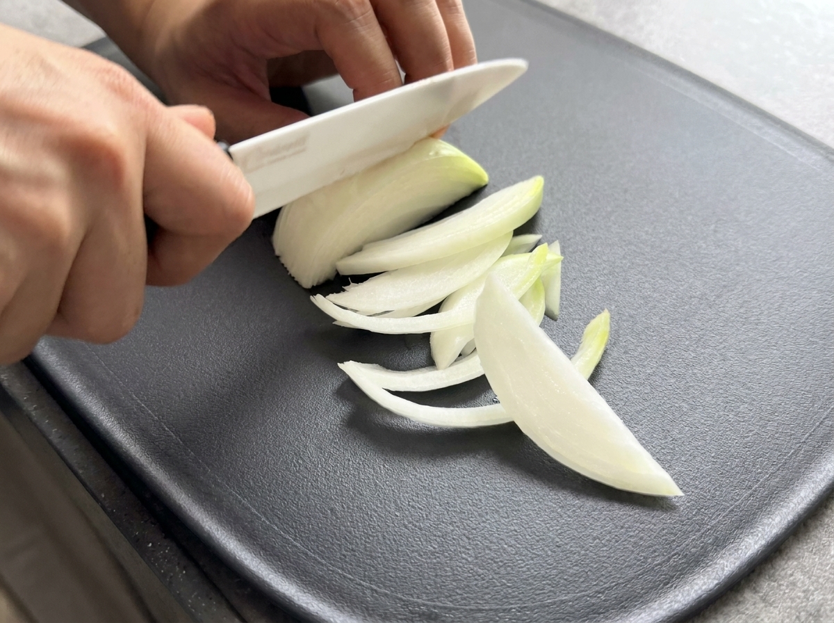 A close-up of a knife slicing a half-onion into thin, uniform strips on a dark surface.