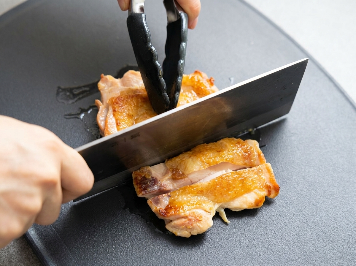 Browned chicken thigh being sliced into pieces on a dark cutting board with a cleaver.