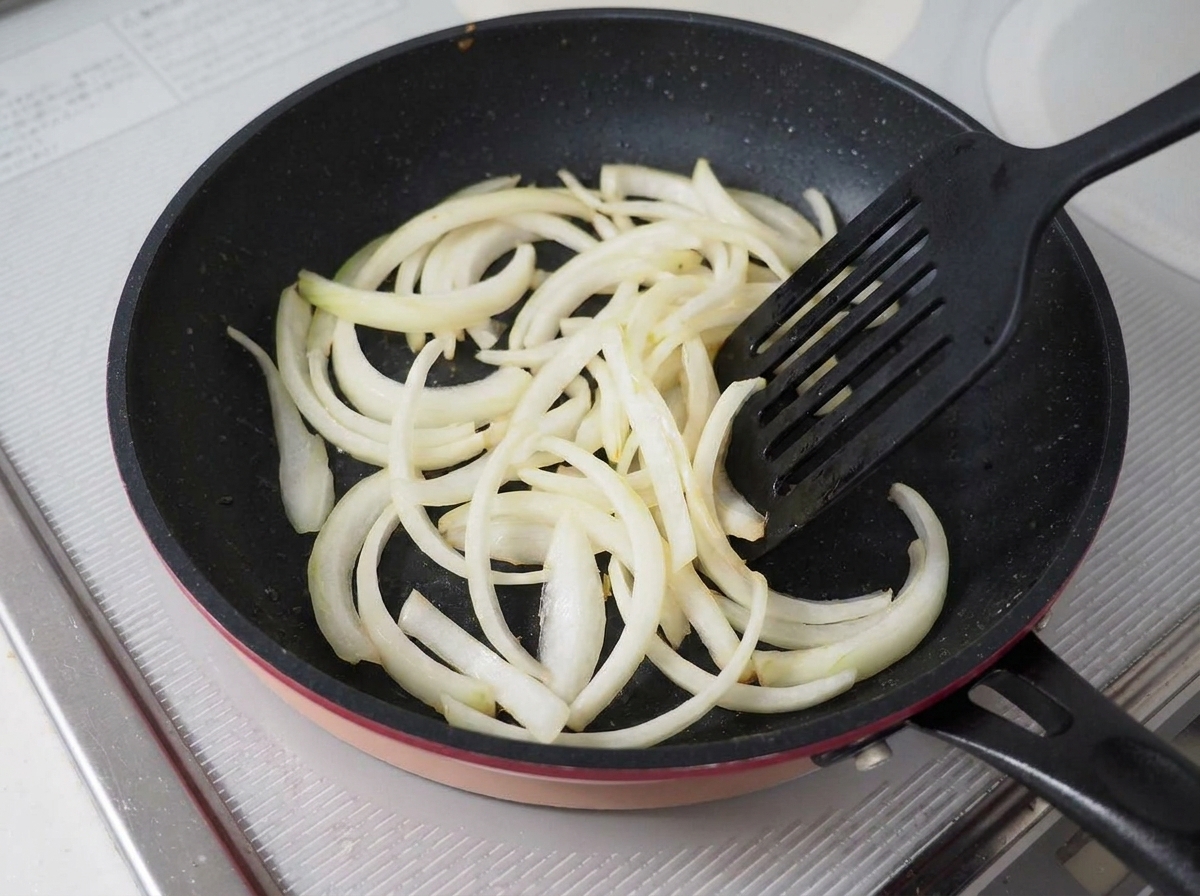 Sliced onions being stir-fried in a dark non-stick pan with a black spatula.