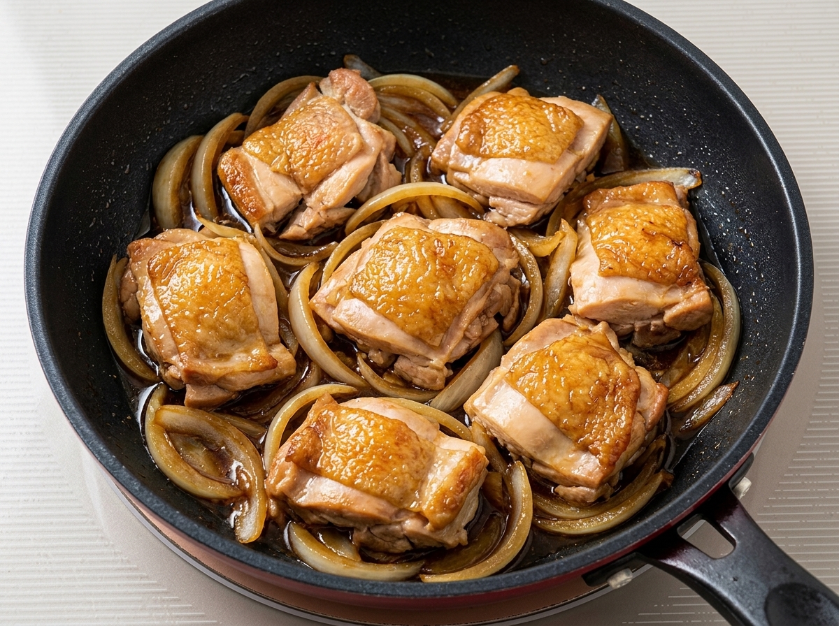Pan-fried chicken pieces simmering with onions and dark sauce in a frying pan.