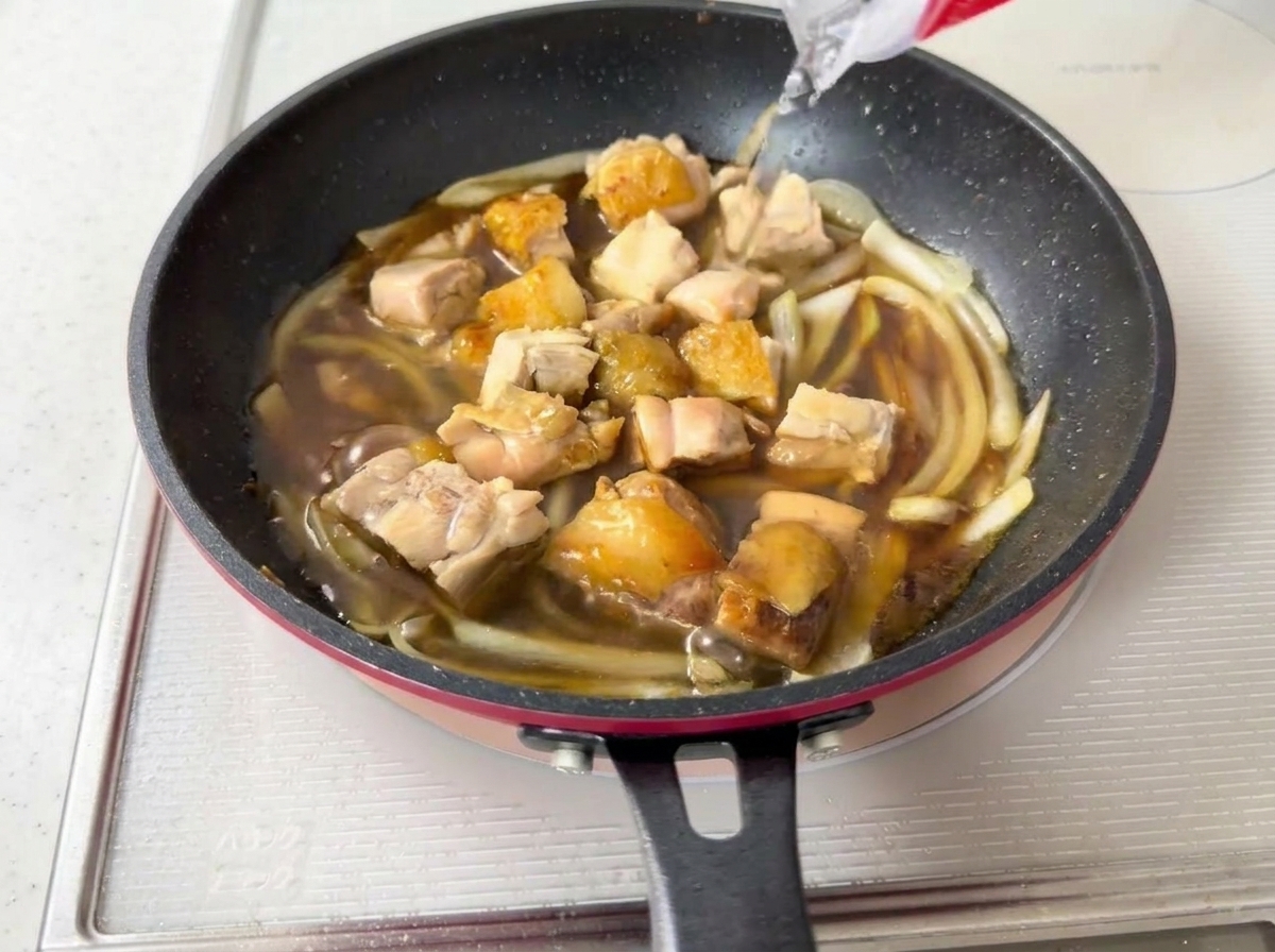 Water being poured into a pan containing simmering chicken, onions, and dark sauce.