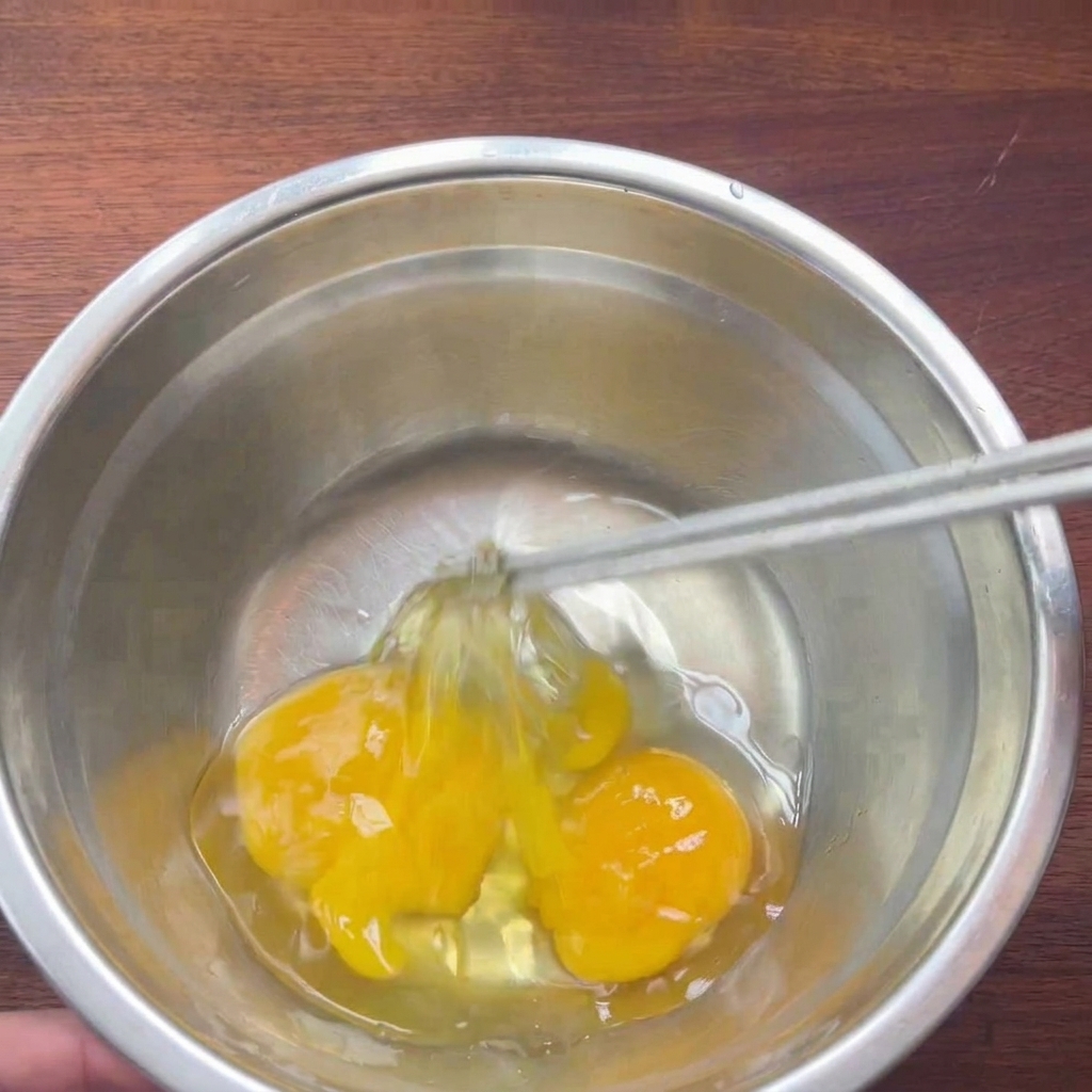Two raw eggs in a metal bowl being gently beaten with chopsticks.