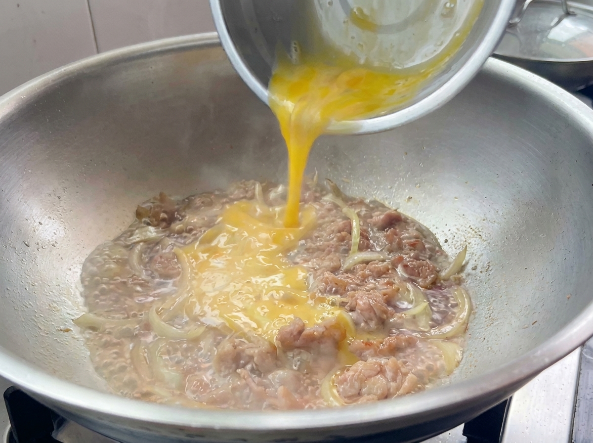 Beaten egg mixture being drizzled over cooked pork and onions in a frying pan.