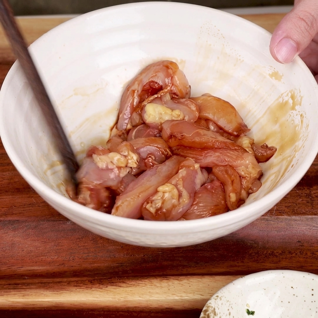 A pair of wooden chopsticks mixing marinated chicken pieces in a white bowl.