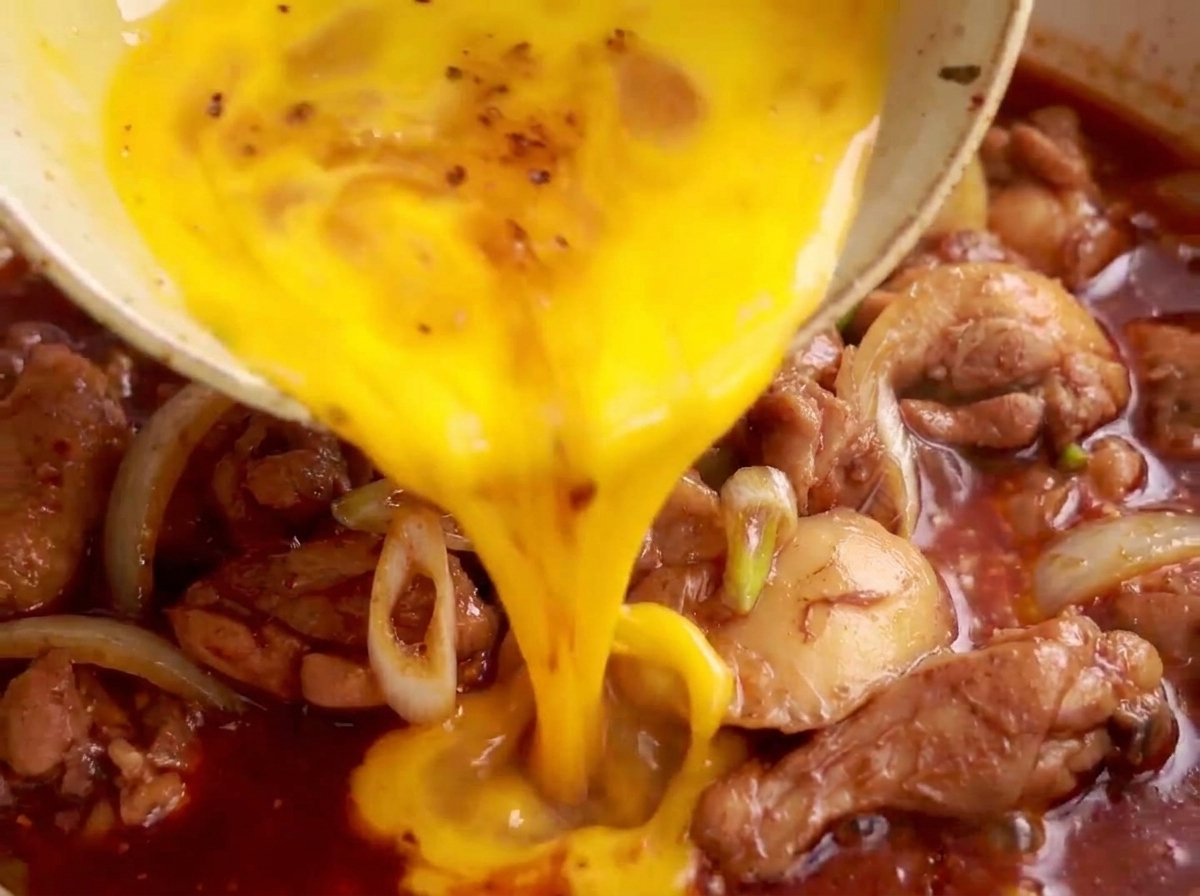 Beaten egg mixture being poured over simmering chicken and leeks in a pan.