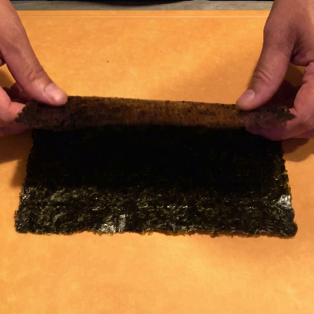A close-up view of hands using kitchen scissors to snip a dark nori sheet into thin, uniform strips over a bowl.