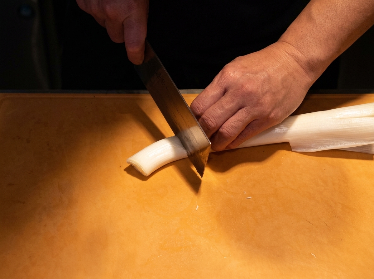 The white stalk of a Japanese long onion being thinly sliced on a wooden cutting board.