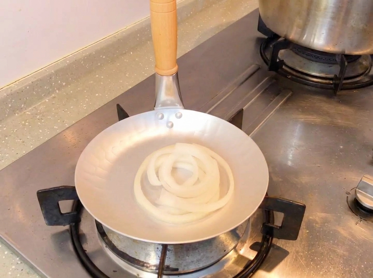 Dark soy sauce mixture being poured from a spoon into a skillet containing raw white onion rings.