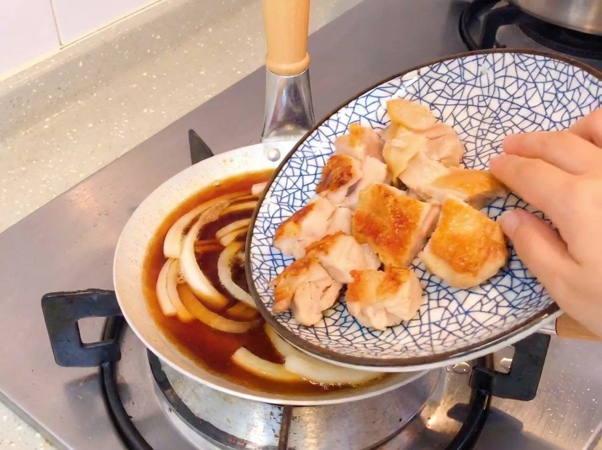 Slices of crispy pan-fried chicken being added to a skillet with simmering onions and soy-based sauce.