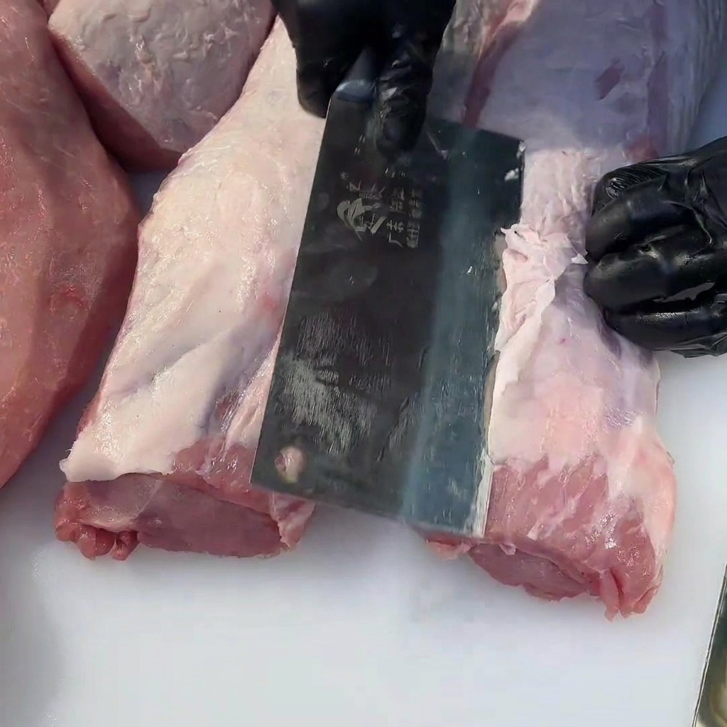 A close-up view showing a meat cleaver trimming excess fat off a raw piece of pork tenderloin.
