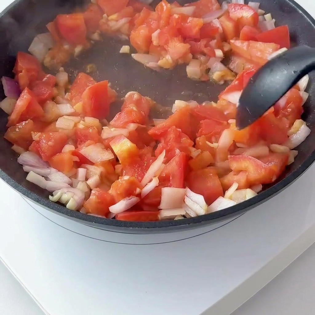 Diced tomatoes and red onion sautéing in a pan with tomato paste being mixed in.