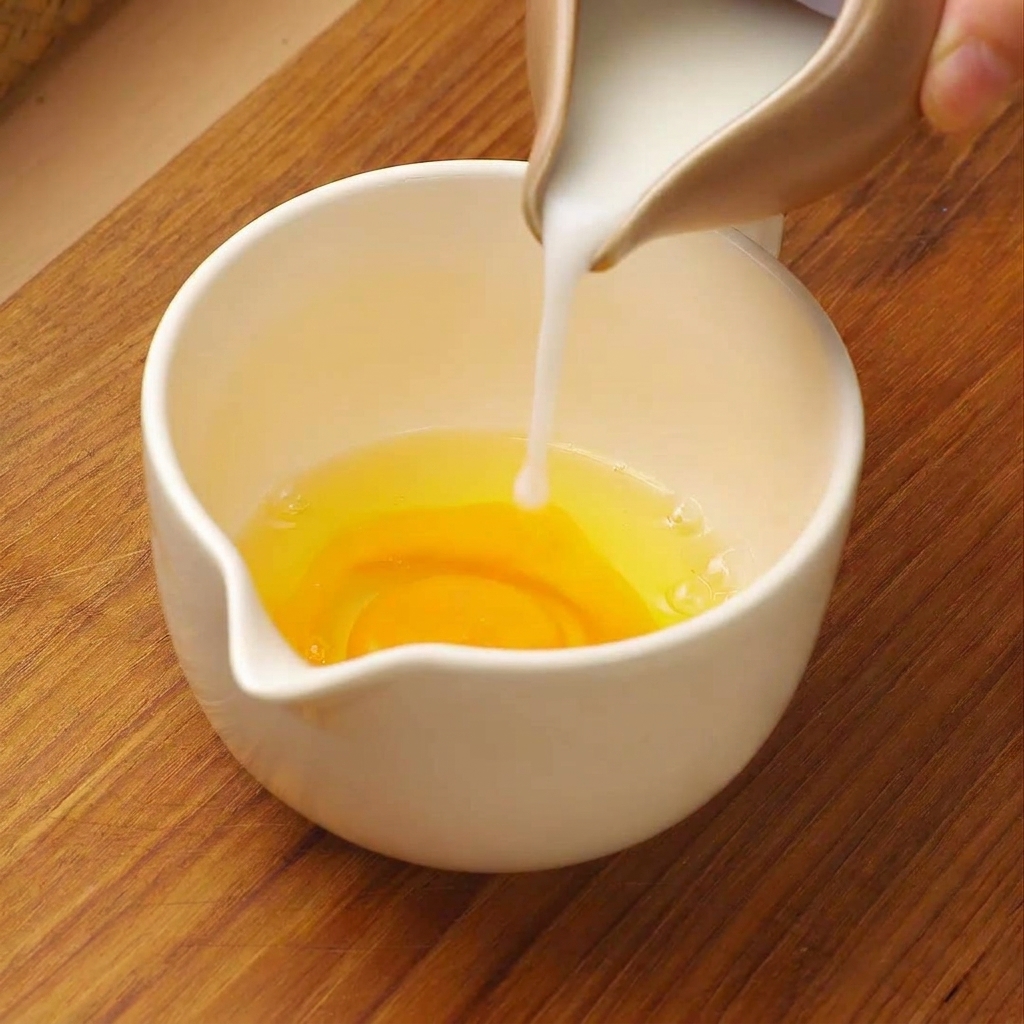 White milk being poured from a small pitcher into a white measuring cup containing raw cracked eggs.