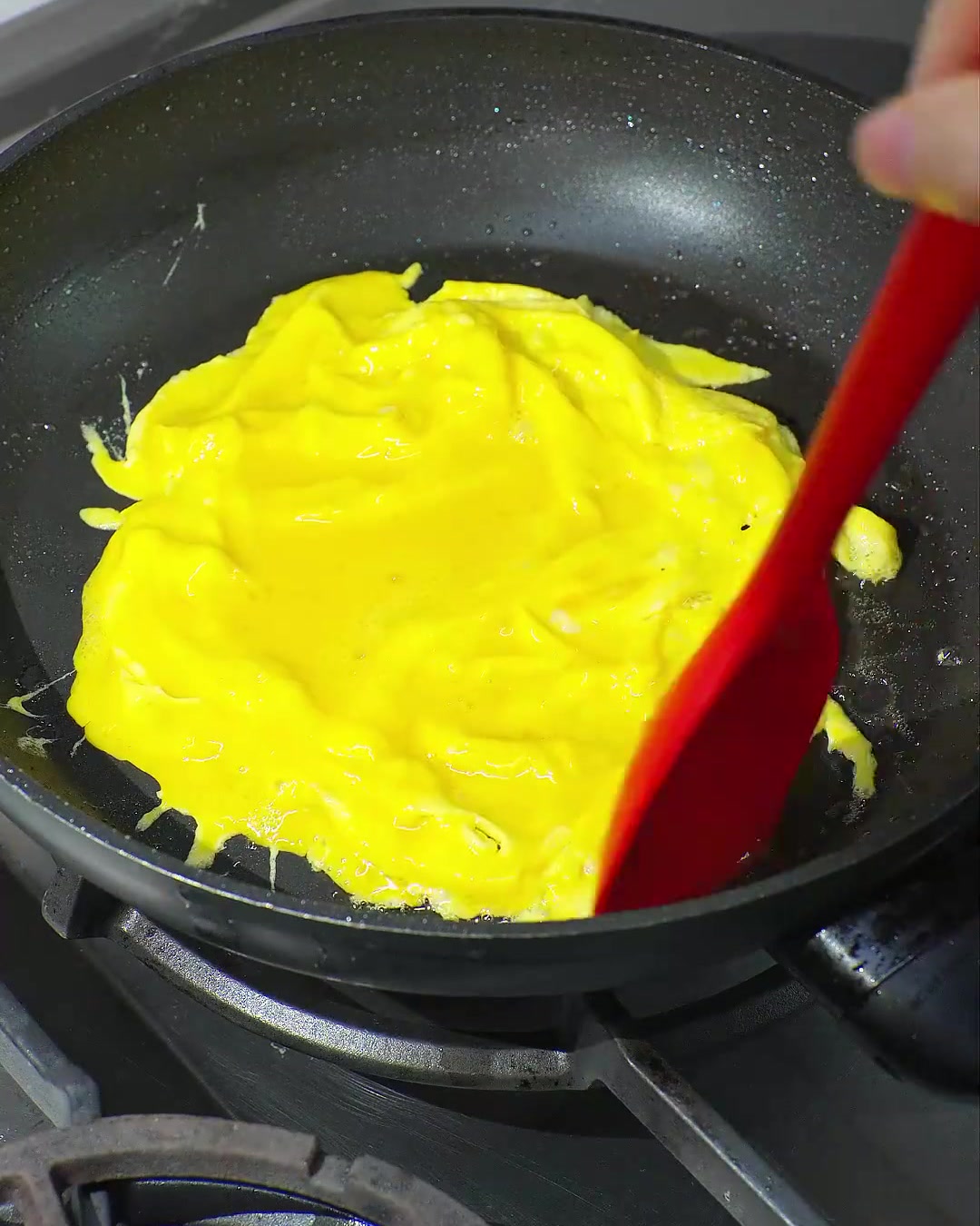 A red spatula folding a bright yellow, partially cooked scrambled egg in a black frying pan.