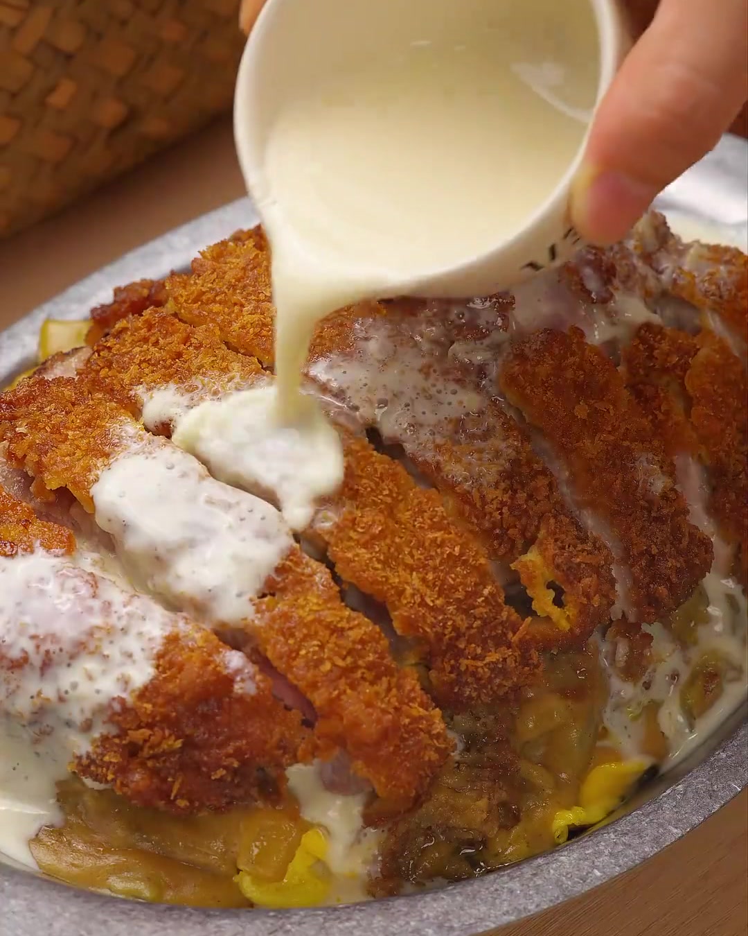 A close-up shot of a creamy white cheese and coconut sauce being poured over golden-brown sliced pork chops, garnished with fresh green parsley.
