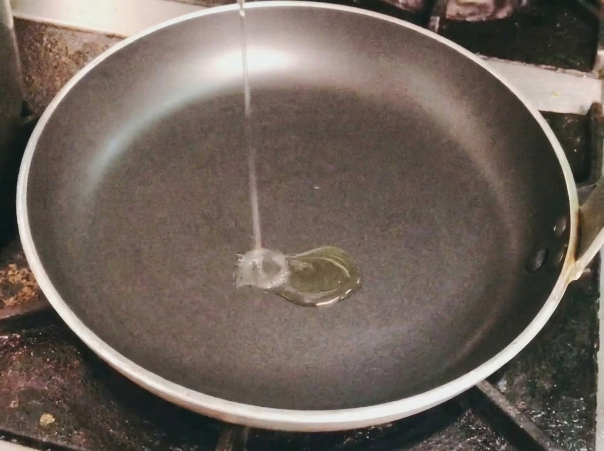A thin stream of cooking oil being poured into an empty black frying pan on a stovetop.