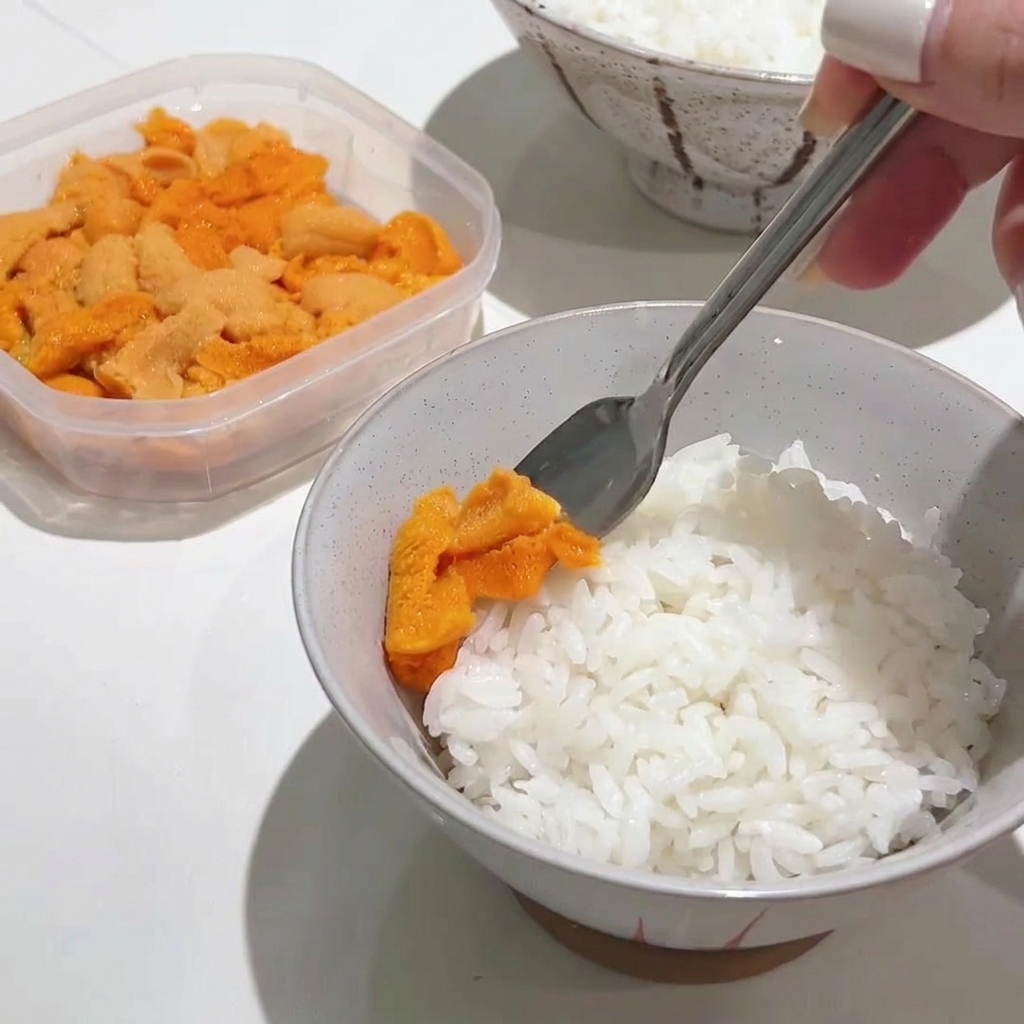 A metal spoon gently placing a generous portion of bright orange sea urchin on top of a bowl of hot white rice.