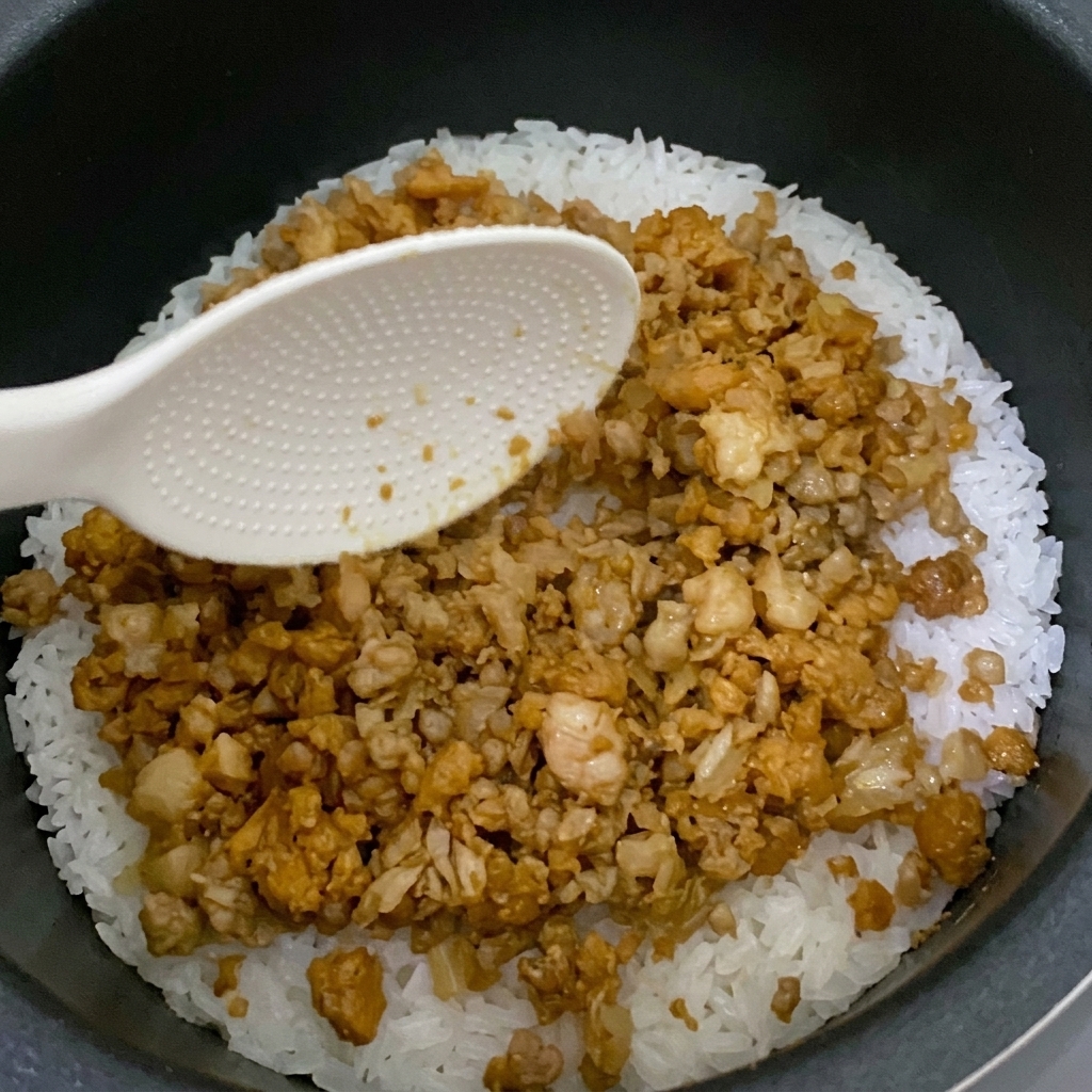 The finished stir-fried mixture being spread over the rice in a rice cooker.