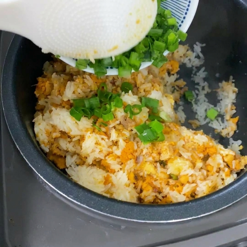 A white spoon pouring freshly chopped green scallions onto yellow-tinted cooked rice inside a rice cooker pot.