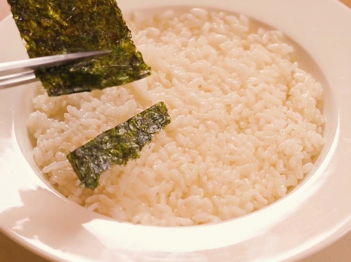 Strips of roasted seaweed being carefully layered across the bed of seasoned rice using chopsticks.