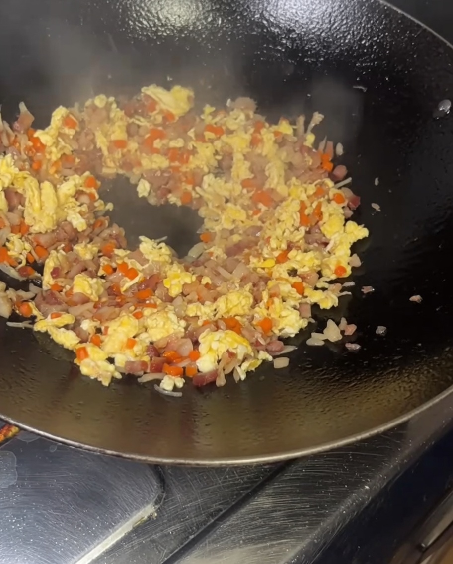 Emptying a bowl of yellow, egg-coated rice grains into a smoking hot wok filled with stir-fried ingredients.