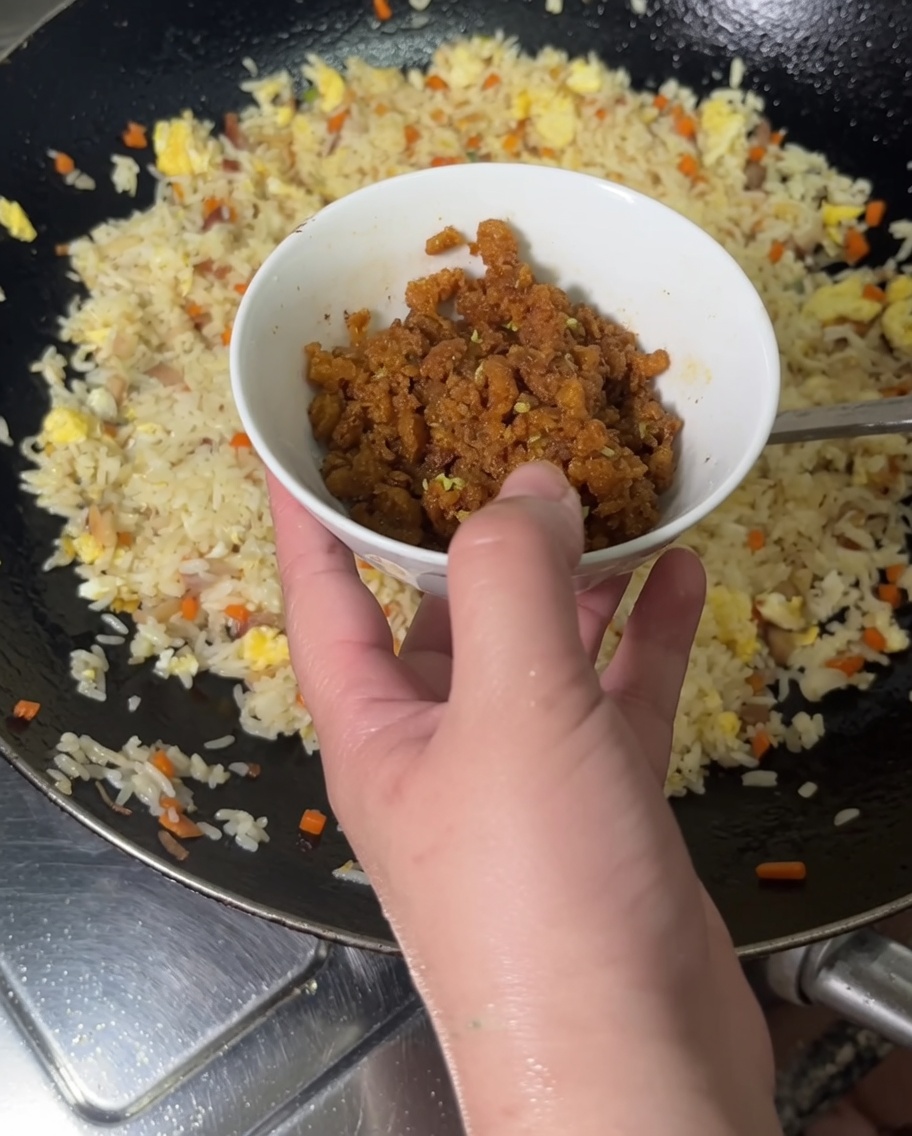 A hand pouring cooked bright orange sea urchin roe from a small bowl into a wok filled with fried rice.