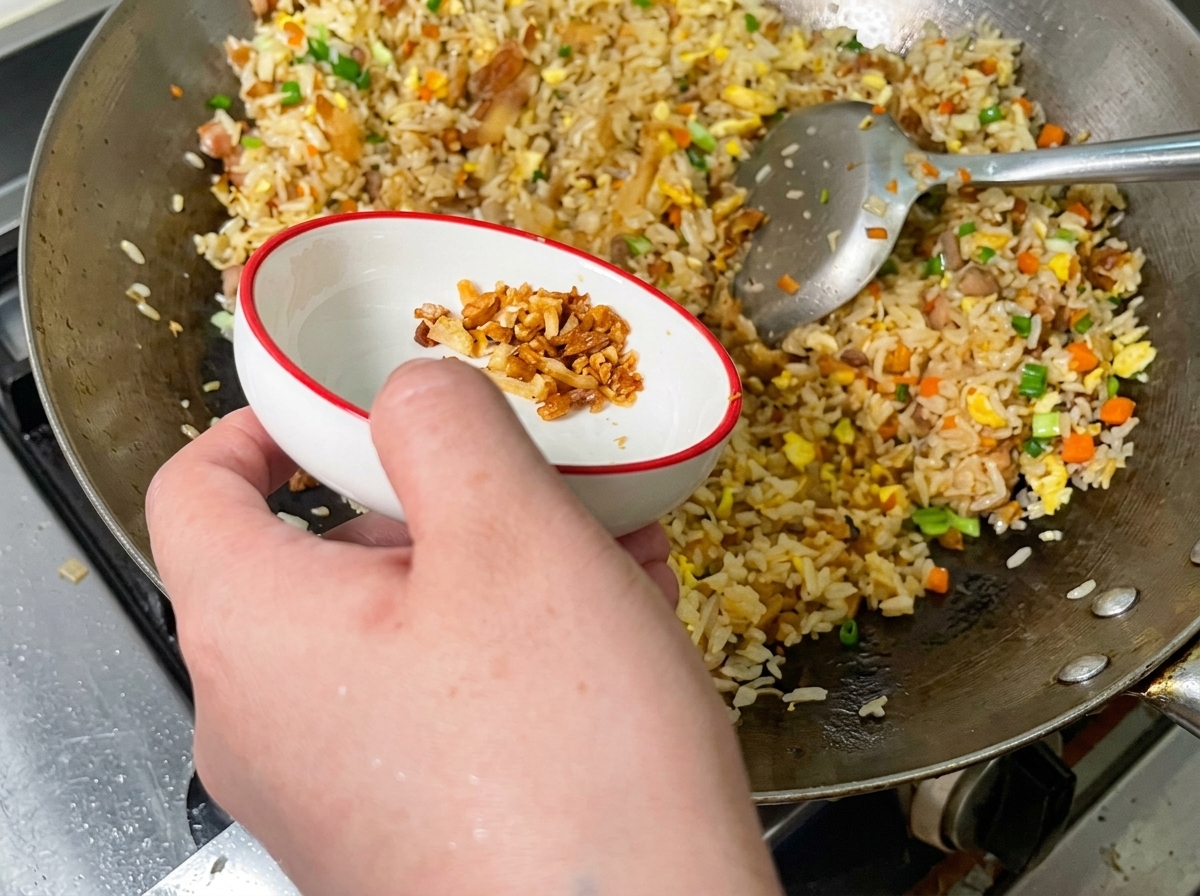 Crispy golden pork cracklings being scattered over a colorful batch of fried rice in a wok.