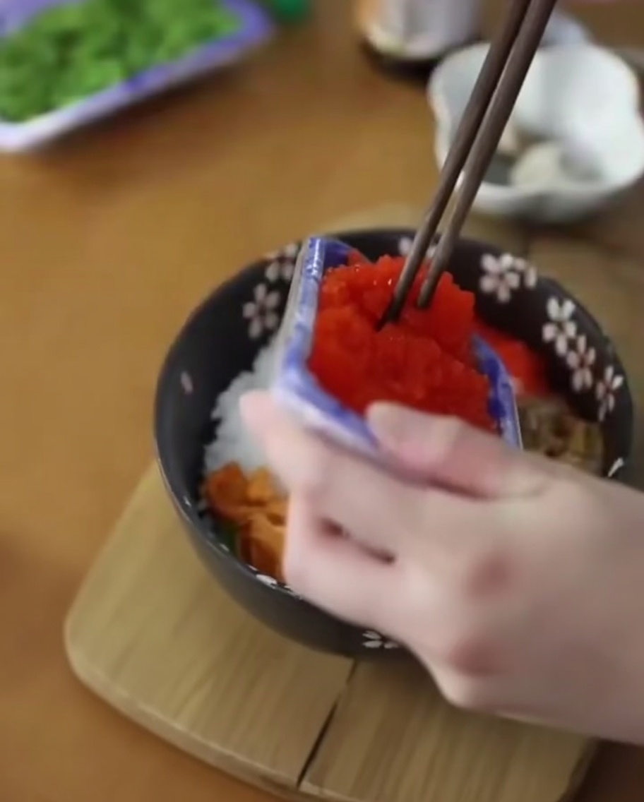 Chopsticks adding orange flying fish roe to a rice bowl already containing sea urchin.