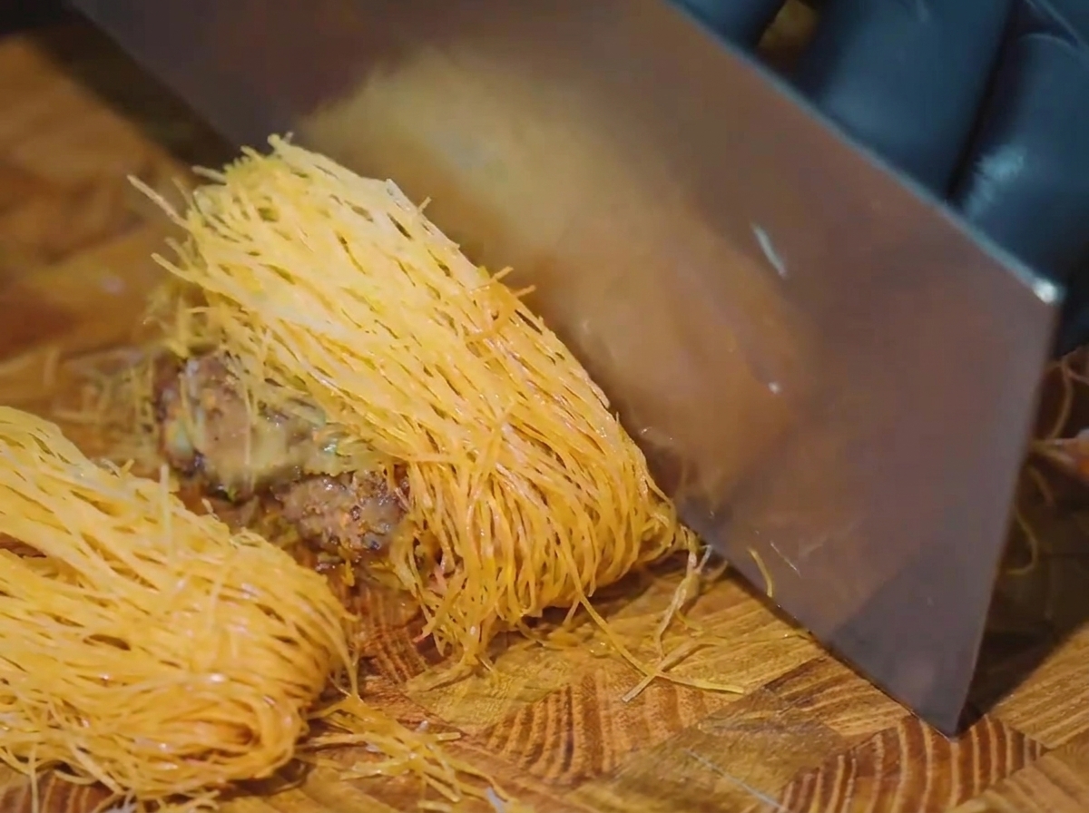 A large cleaver slicing straight down into a golden brown, thread-wrapped beef log on a wooden cutting board.