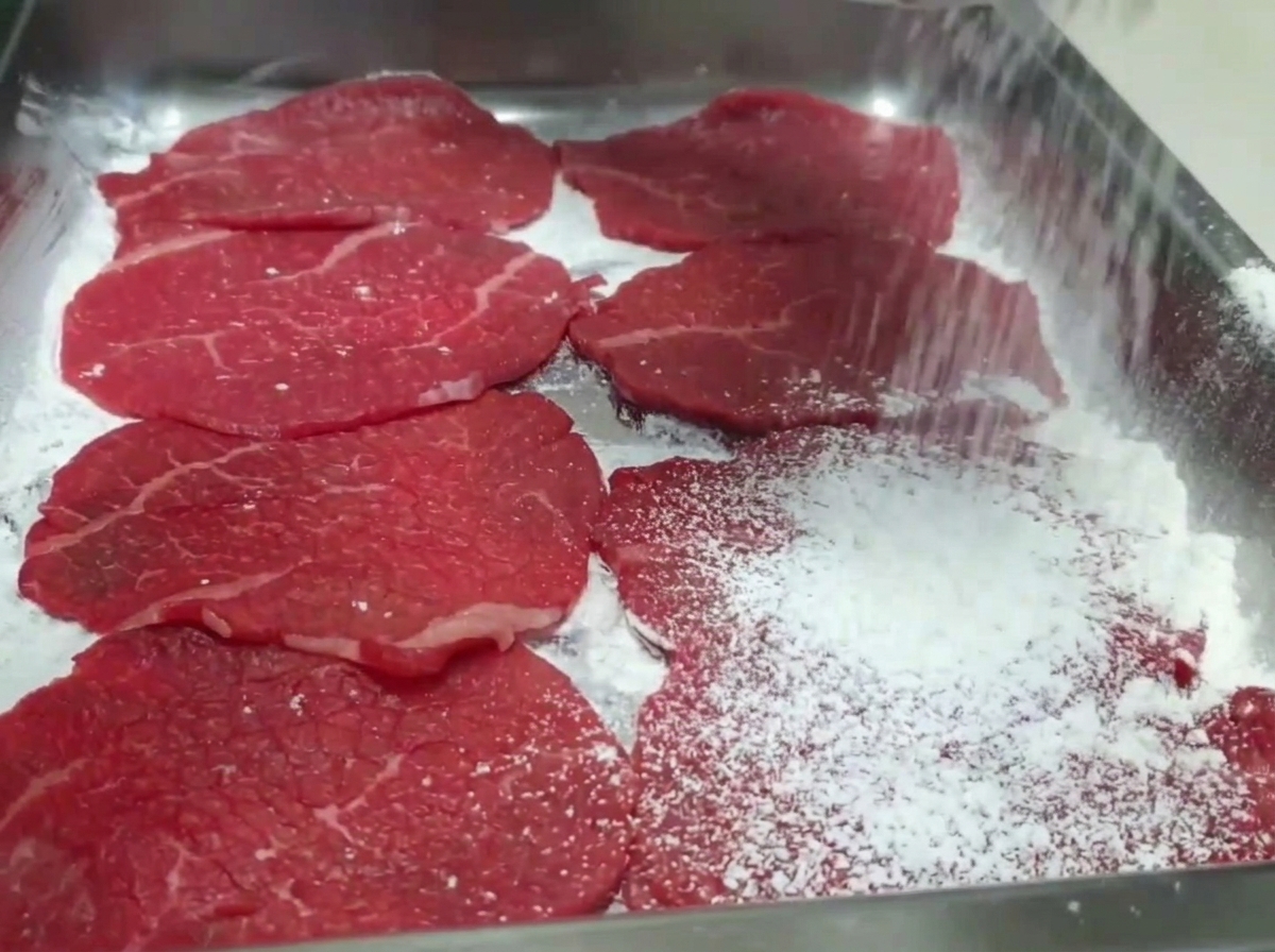 Hand dusting white flour over raw beef slices arranged in a tray.