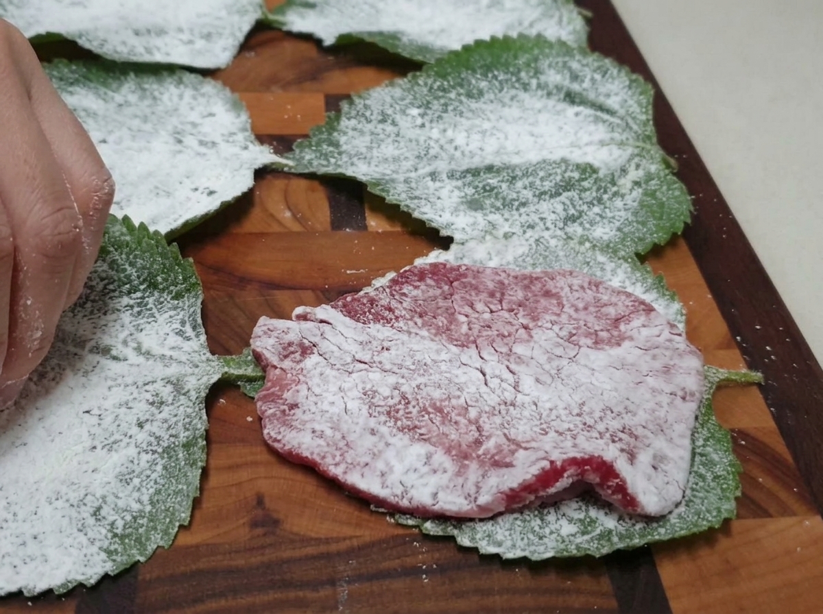 A floured beef slice being positioned on top of a floured green perilla leaf on a wooden cutting board.