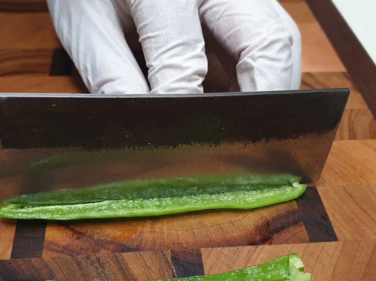 A chefs knife finely chopping a green chili pepper on a wooden cutting board.
