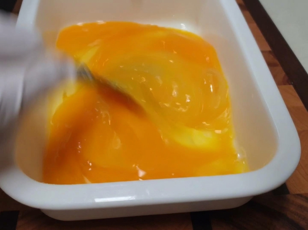 A whisk blending bright yellow egg yolks and whites inside a white rectangular tray.