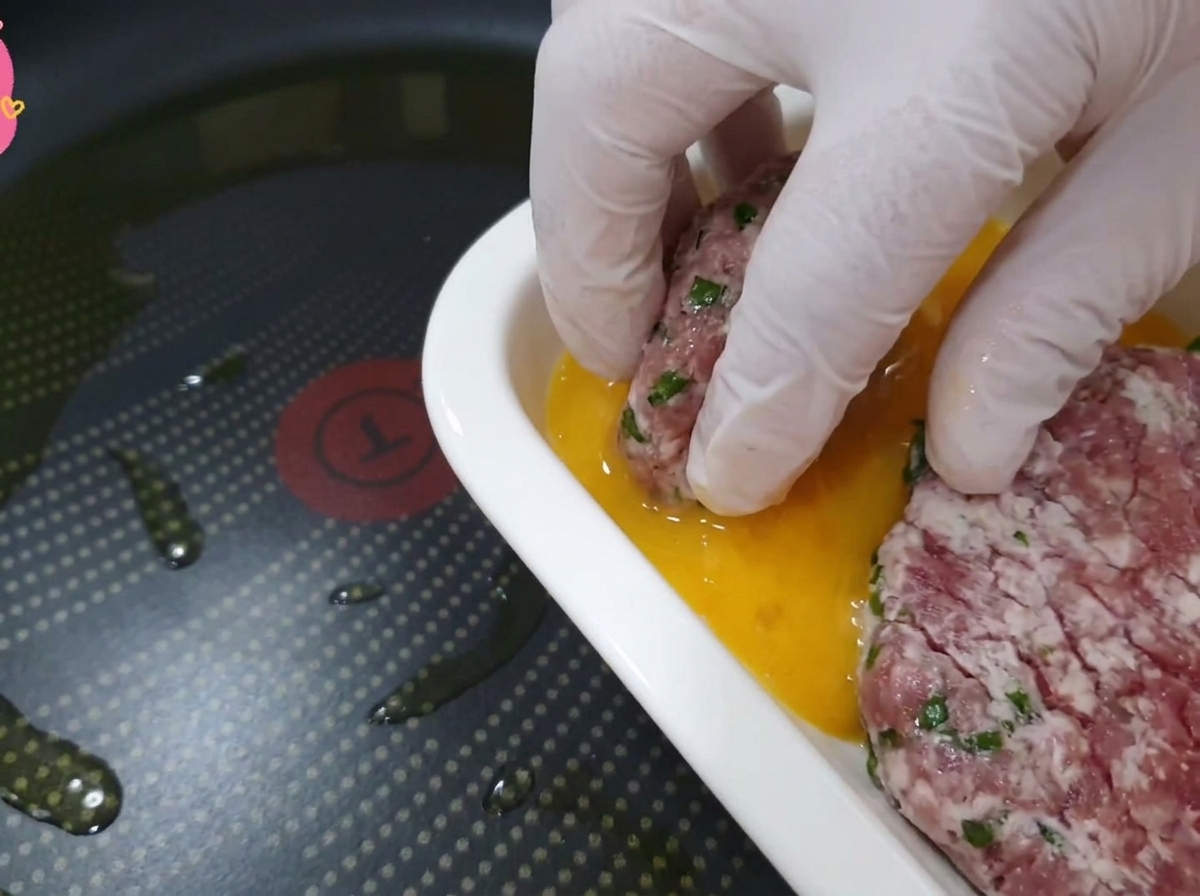 Hand in a clear glove dipping a piece of beef and perilla leaf into a dish of golden egg wash.