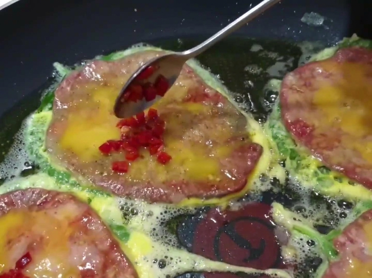 Close-up of a spoon adding diced green and red chili peppers to the top of cooking beef pancakes in a pan.