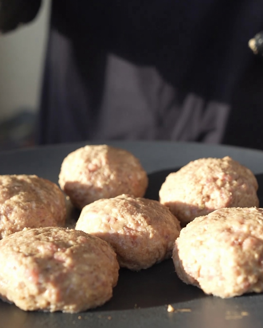 Round meat patties cooking on a flat black cast-iron pan over low heat.