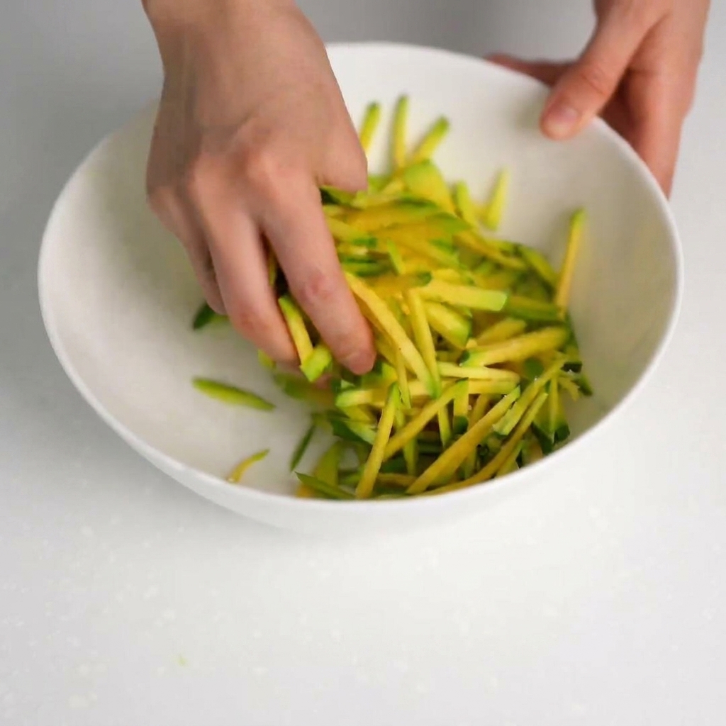Hands pressing down on salted zucchini strips to drain liquid into a clean bowl.