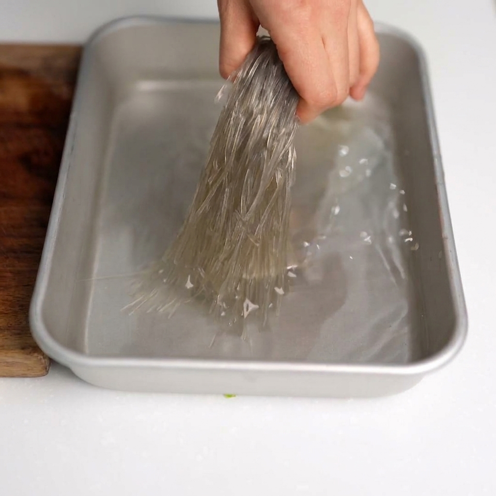 Kitchen scissors cutting soaked glass noodles into small segments inside a mixing bowl.