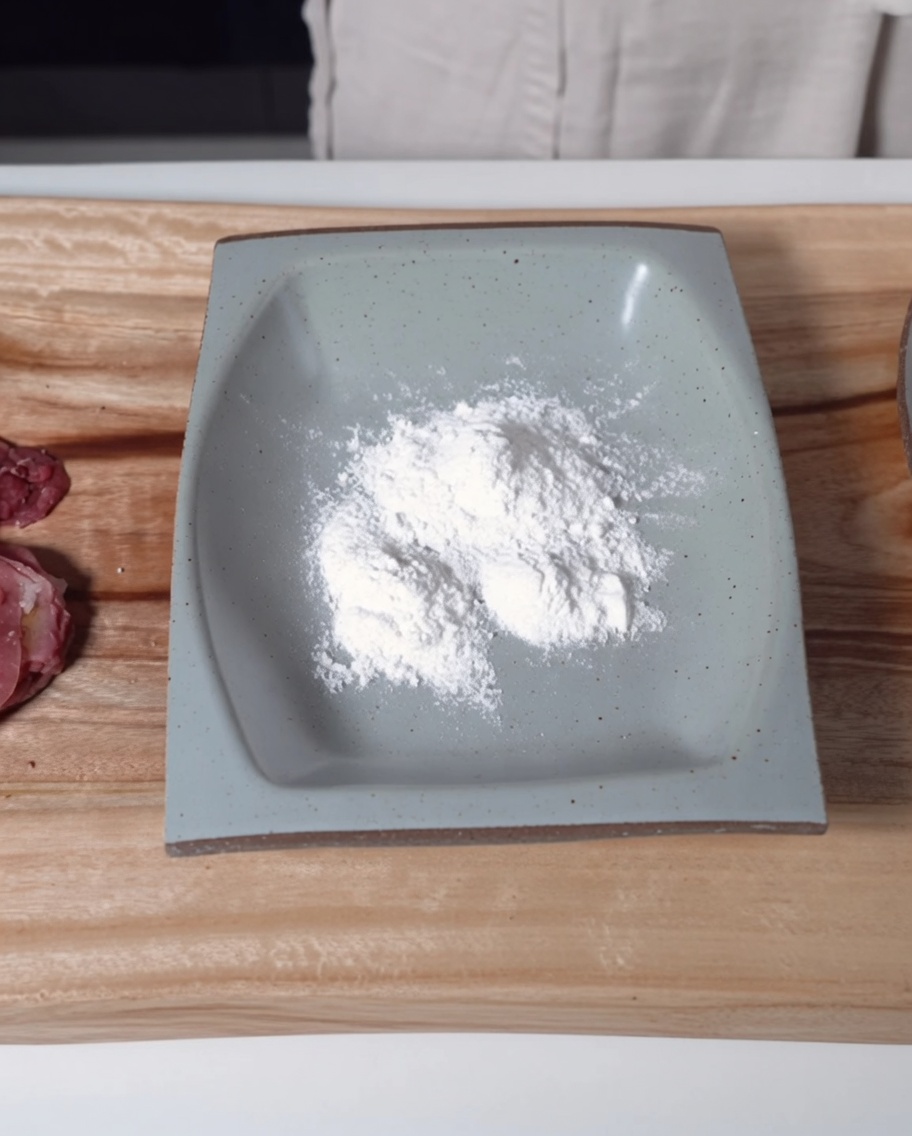 A shallow grey rectangular dish filled with fine white glutinous rice flour next to seasoned raw beef slices.