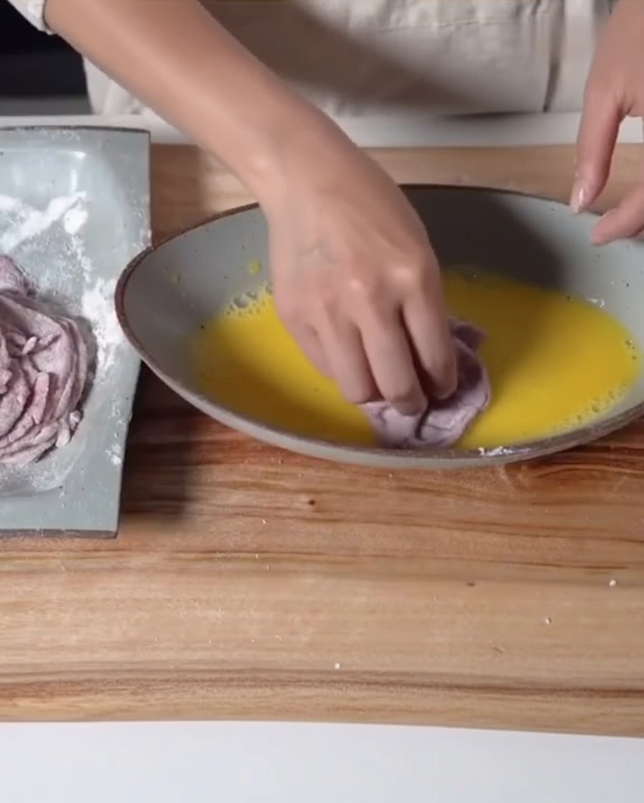 A hand dipping a flour-coated piece of beef into a shallow dish filled with bright yellow beaten eggs.